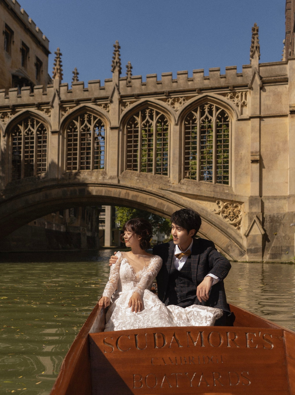 A bride in a white lace wedding gown and a groom in a dark suit sit together in a wooden boat on a canal, with a historic stone bridge and buildings in the background under a blue sky.