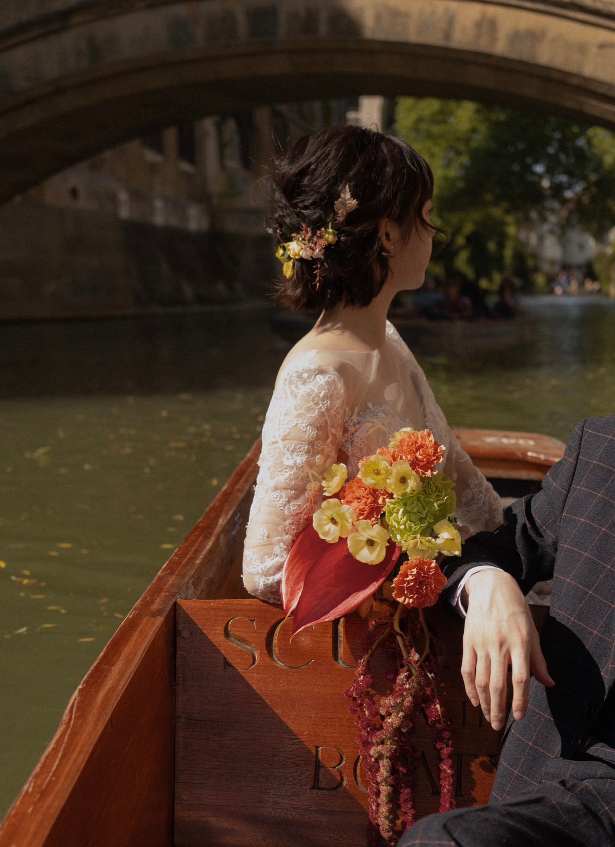 A woman in a white lace wedding dress sits in a wooden boat on a canal, holding a bouquet of yellow and orange flowers. A man in a dark plaid suit is seated beside her, with a stone bridge and green water in the background.
