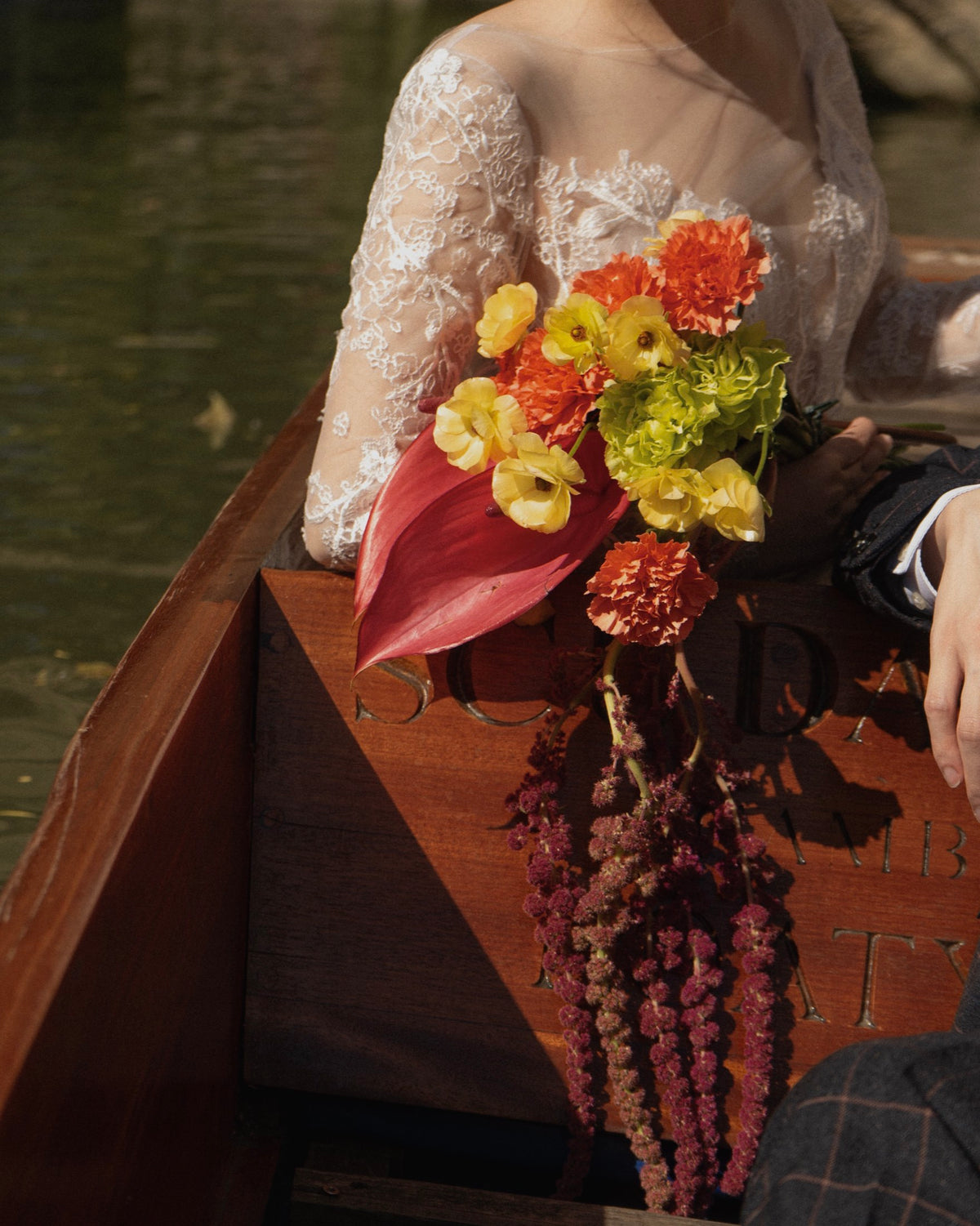 A bride in a white lace gown sits in a wooden boat holding a vibrant bouquet of yellow, orange, and red flowers.