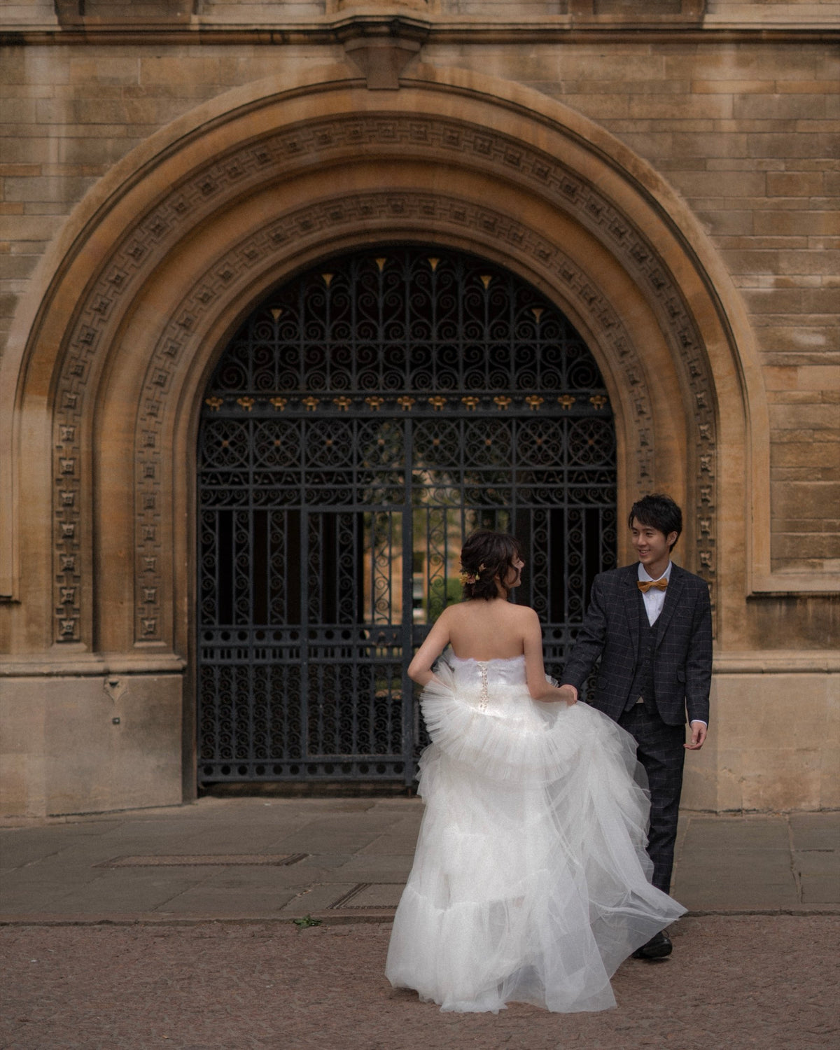 A bride in a strapless white tulle wedding gown and a groom in a dark suit stand in front of a large stone archway with an iron gate.