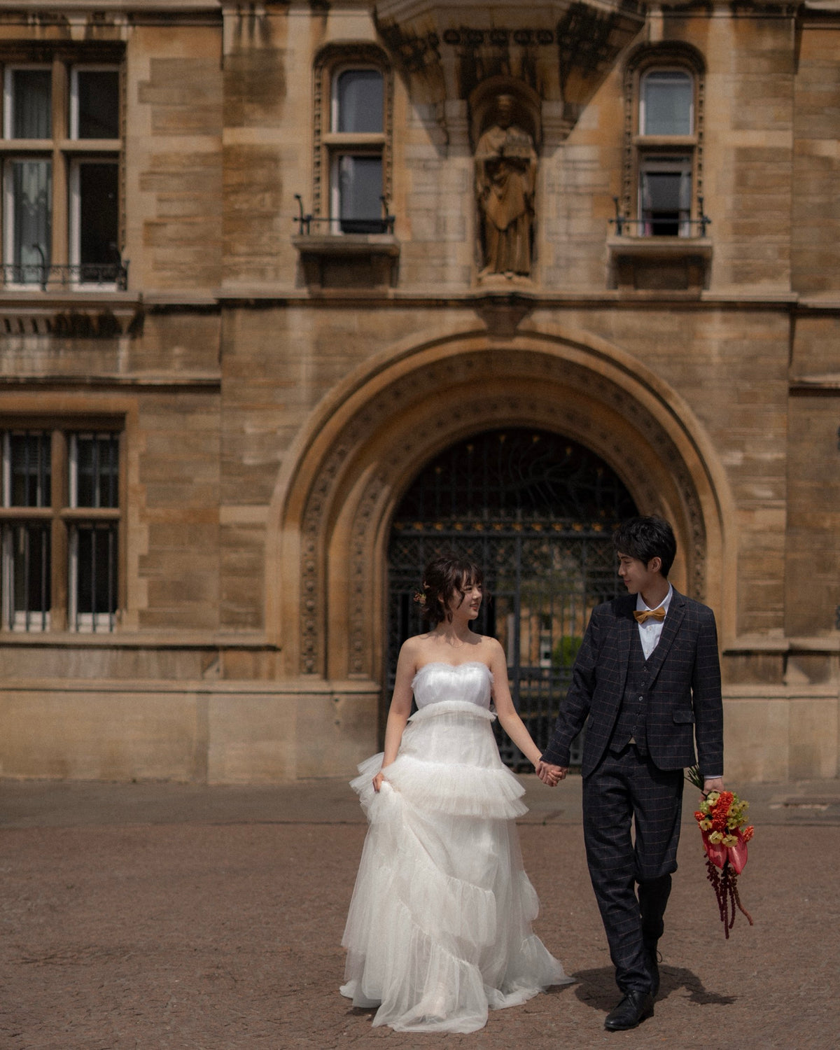 A bride in a white strapless tulle wedding gown and a groom in a dark gray suit walk hand-in-hand in front of a historic stone building with an arched entrance.
