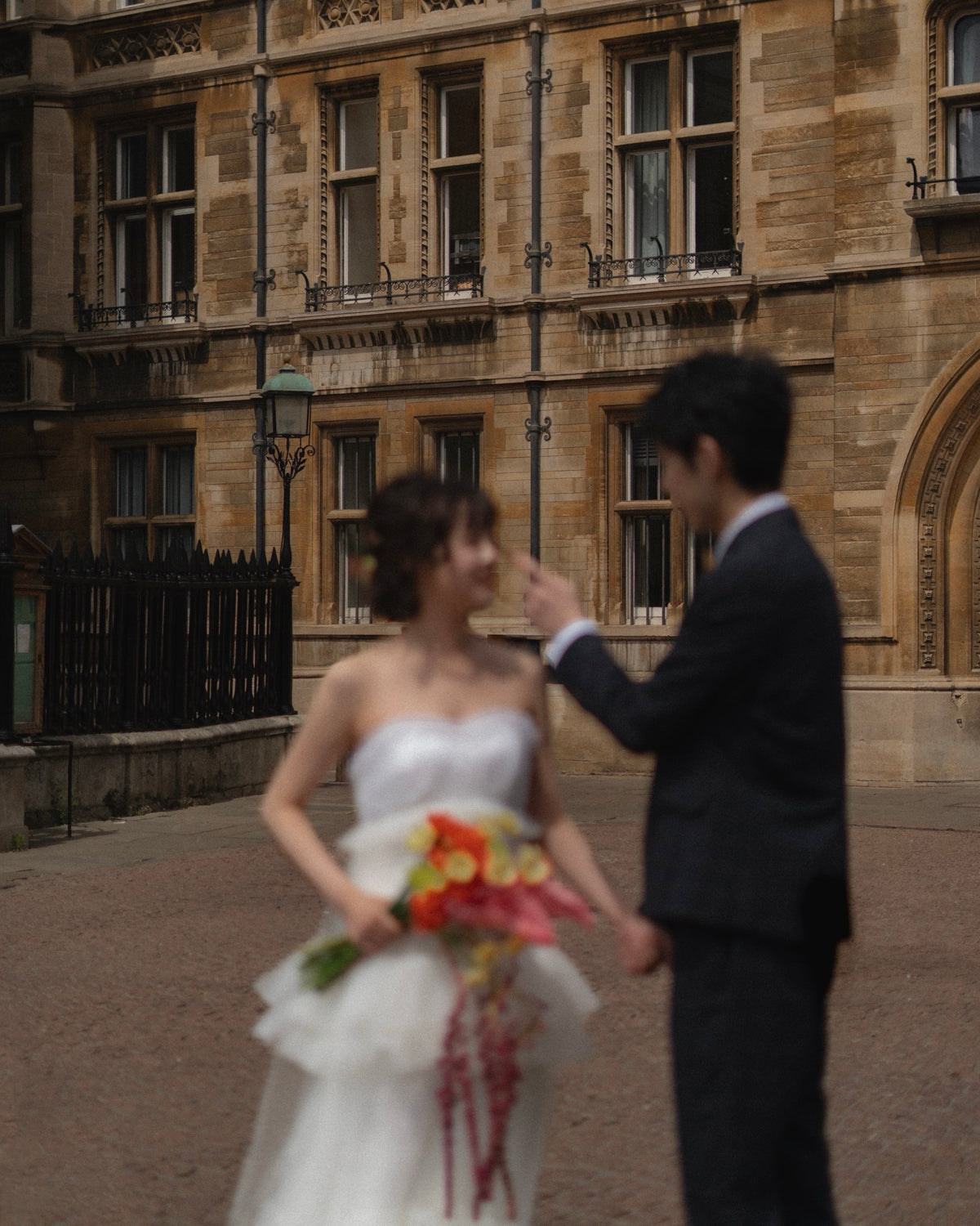 A bride in a white strapless wedding gown holding a colorful bouquet stands in front of a man in a dark suit, with a blurred historic stone building in the background.
