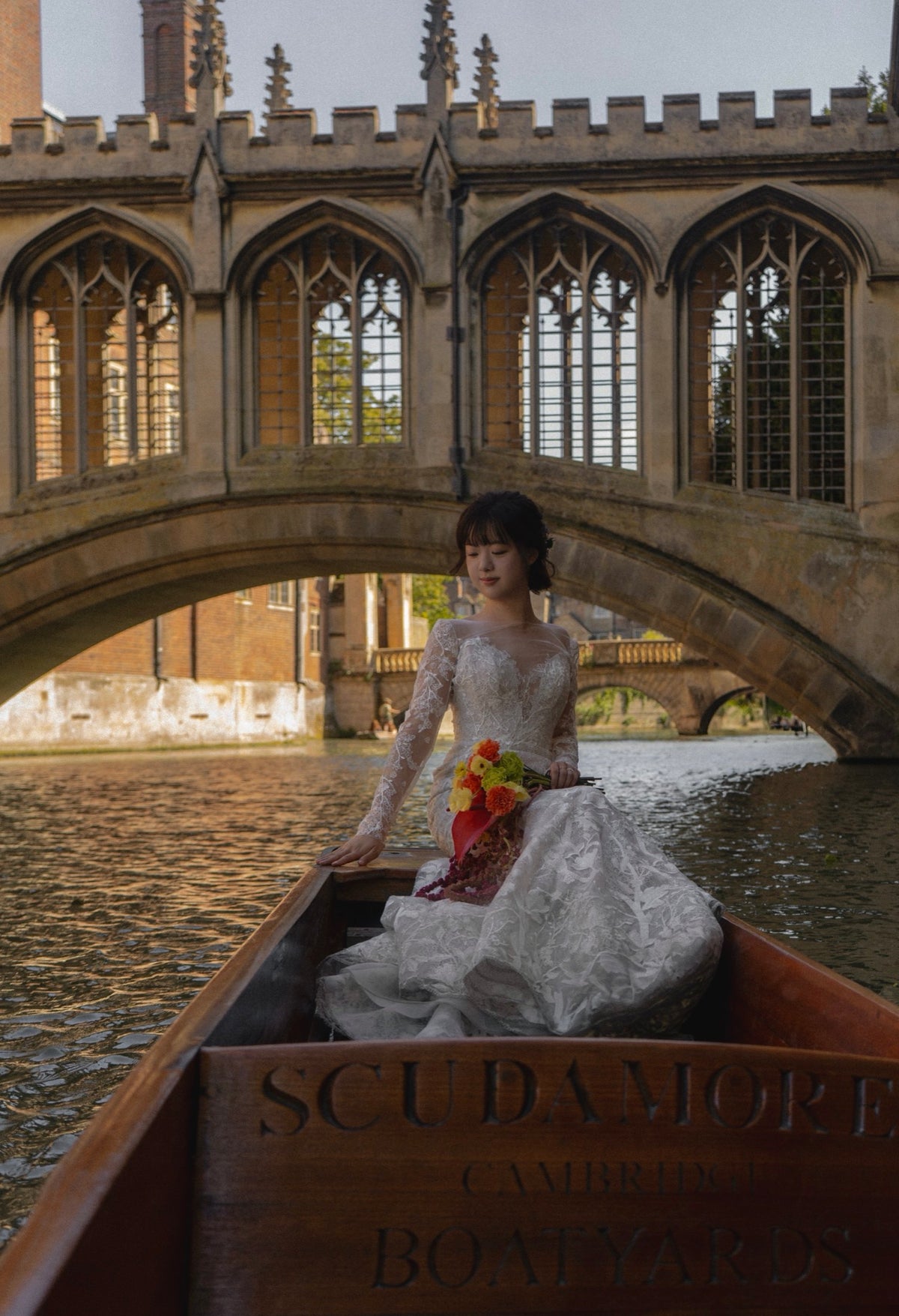 A woman in a white lace wedding gown sits in a wooden boat on a river, with a stone bridge and historic buildings in the background. The boat has 'SCUDAMORE CAMBRIDGE BOATYARDS' carved on the front.