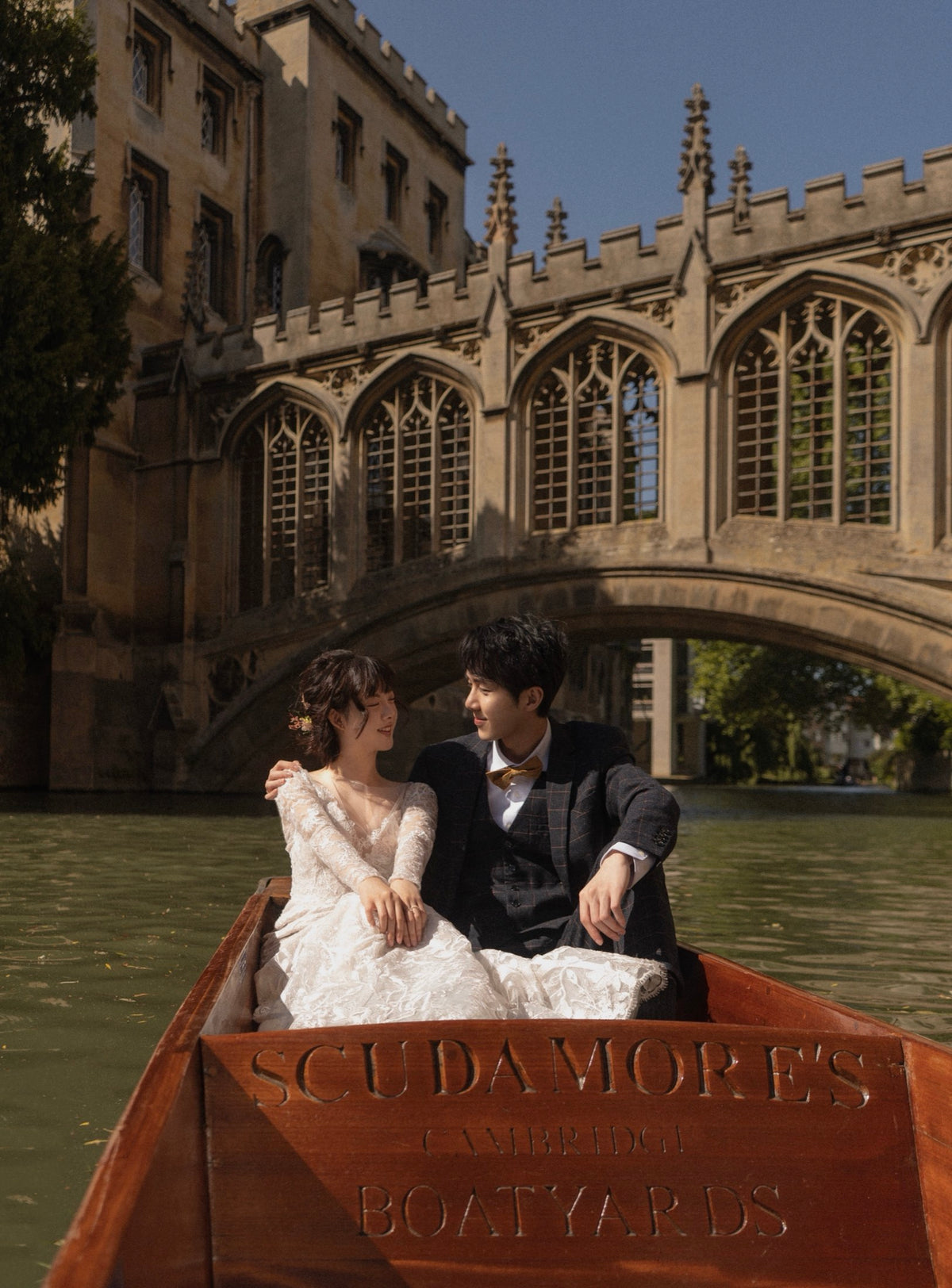 A couple posing in a wooden boat on a river, with the bride in a white lace gown and the groom in a dark suit, set against the backdrop of Cambridge's Stone Bridge and historic buildings.