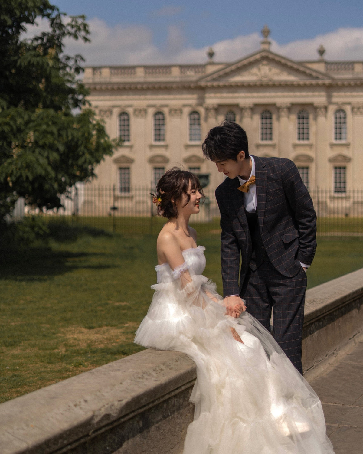 A couple posing outdoors in front of a grand classical building. The woman wears a voluminous off-the-shoulder white wedding gown with a long train, and the man wears a dark gray plaid suit with a yellow bow tie.