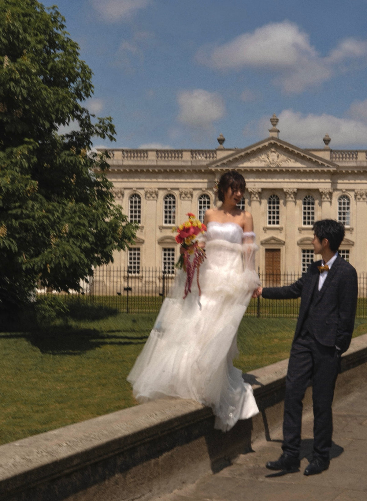A bride in a white off-the-shoulder wedding gown holding a colorful bouquet stands on a stone wall next to a man in a dark suit, with a grand classical building and green lawn in the background under a blue sky.