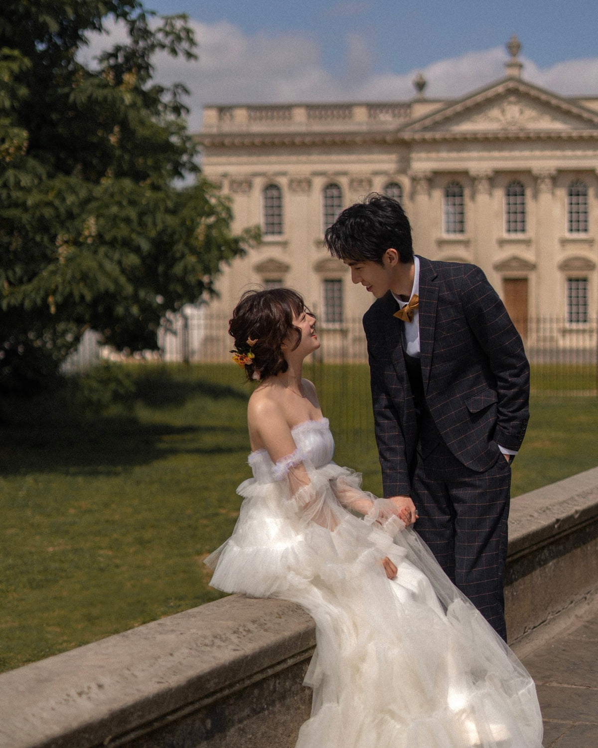 A bride in a white strapless tulle wedding gown and a groom in a dark plaid suit stand together outdoors in front of a grand classical building, with the bride leaning on a stone wall.