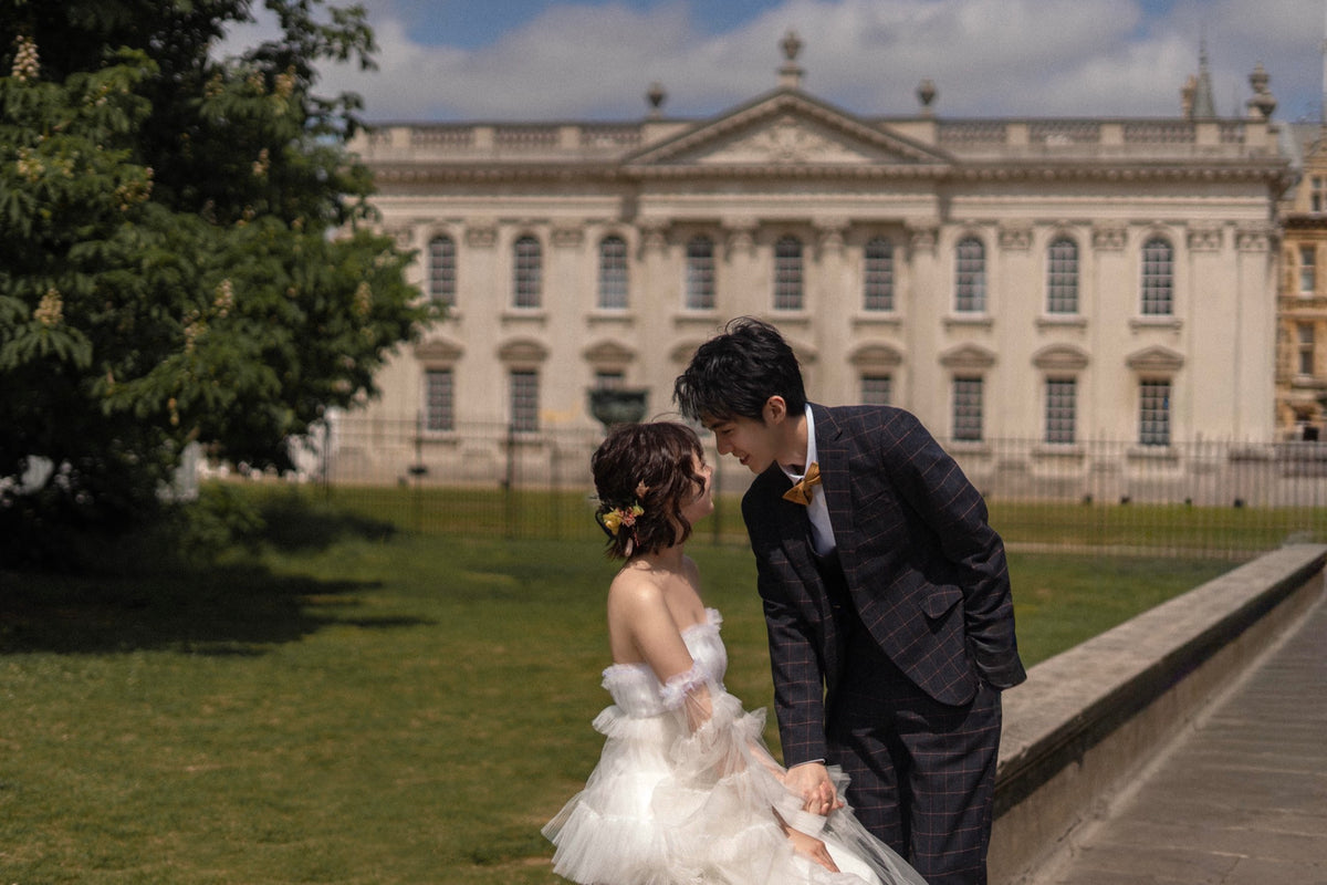A bride in a white tulle wedding dress and a groom in a dark plaid suit stand together outdoors in front of a grand classical building. The bride has a small yellow flower in her hair, and the groom wears a matching yellow boutonniere.
