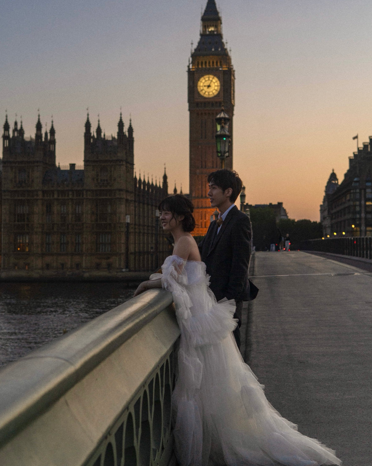 A couple posing on Westminster Bridge in London at sunset, with Big Ben and the Houses of Parliament in the background. The woman is wearing an off-the-shoulder white tulle wedding gown, and the man is wearing a dark suit.