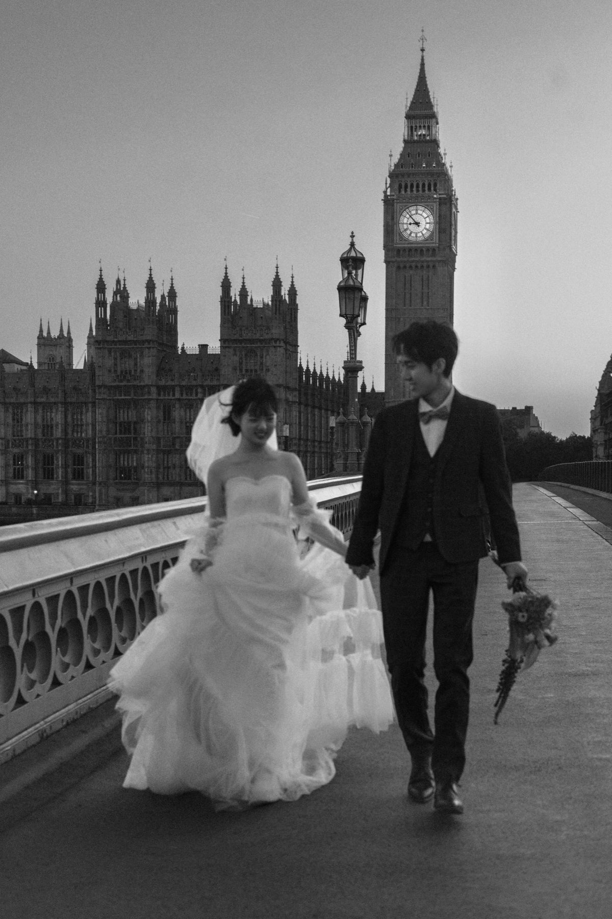Black and white wedding photograph of a bride and groom walking hand in hand on Westminster Bridge, with Big Ben and the Houses of Parliament in the background.