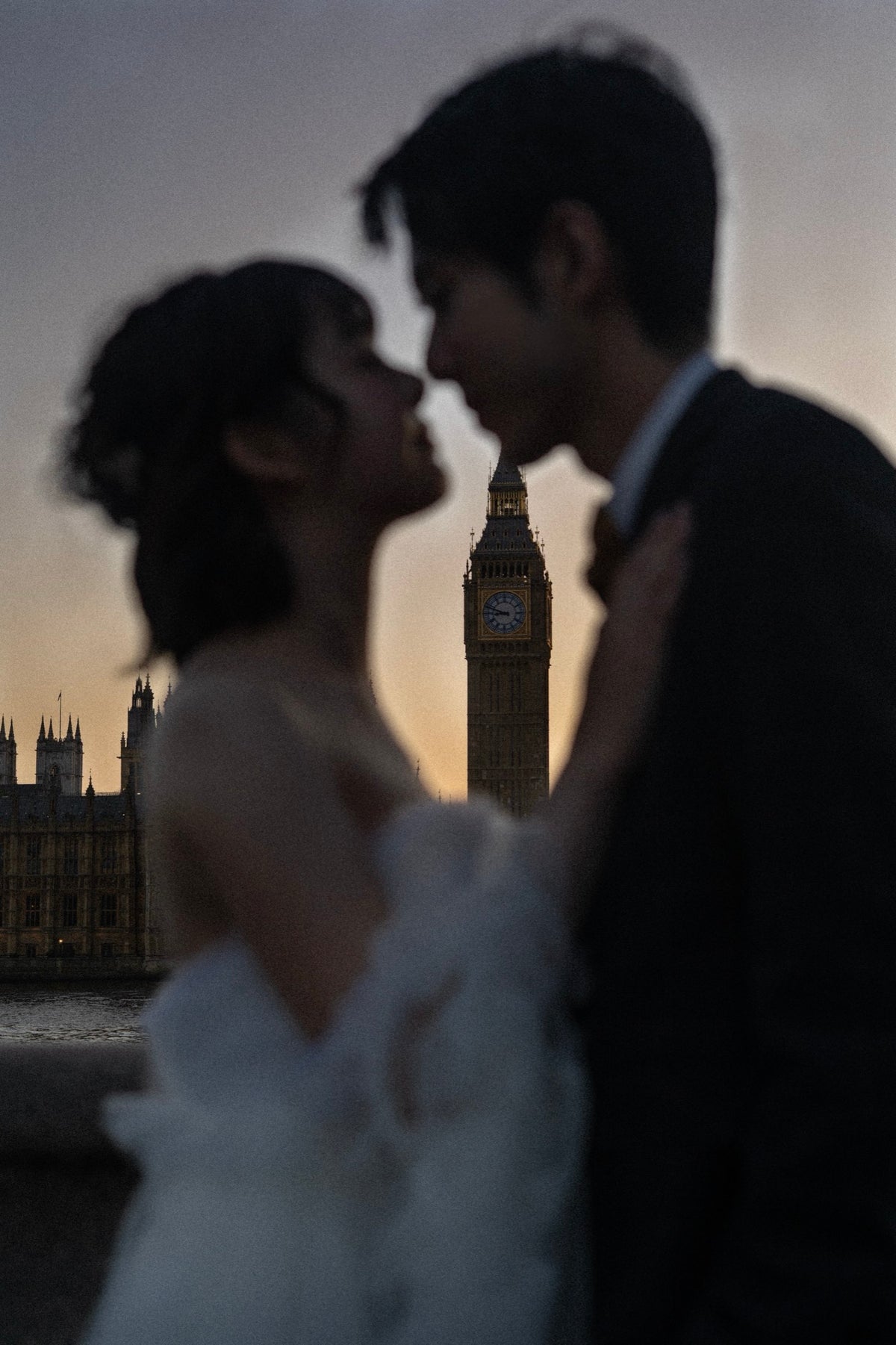 A couple in wedding attire embracing and kissing, with Big Ben and the London Eye silhouetted against a warm sunset sky in the background.