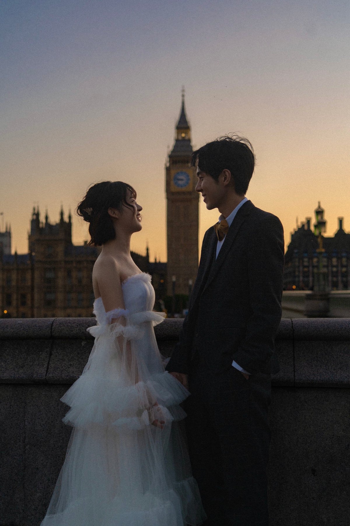 A couple posing together in front of Big Ben and the London Eye at sunset. The woman is wearing a white strapless tulle wedding gown, and the man is wearing a dark suit.
