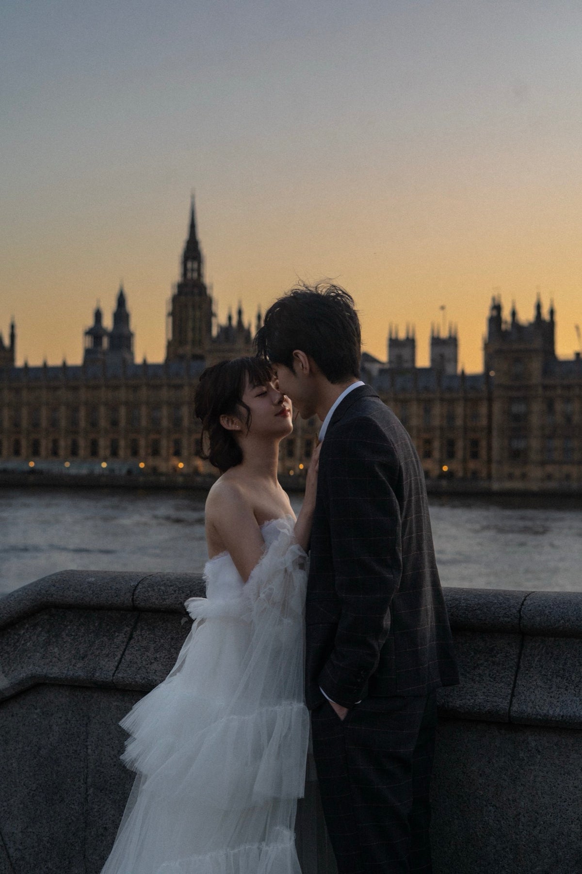 A bride in a white tulle wedding gown and a groom in a dark suit kiss on a stone bridge with the London Palace of Westminster and River Thames visible in the background at sunset.