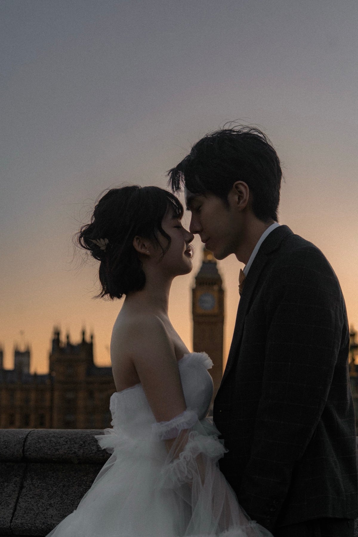 A couple in wedding attire embracing closely at sunset, with a London landmark tower and buildings visible in the background. The woman wears a white strapless wedding dress, and the man wears a dark suit jacket.