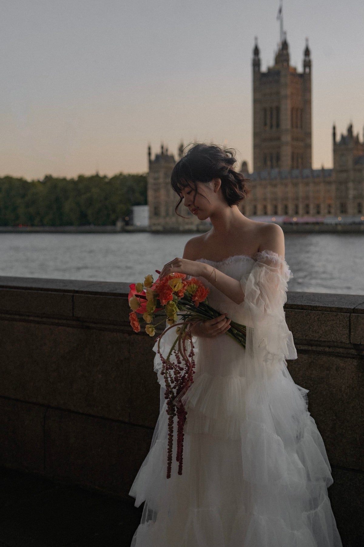 A woman in an off-the-shoulder white tulle wedding gown stands by a stone railing at dusk, holding a bouquet of orange and yellow flowers. The London Palace of Westminster and River Thames are visible in the background.