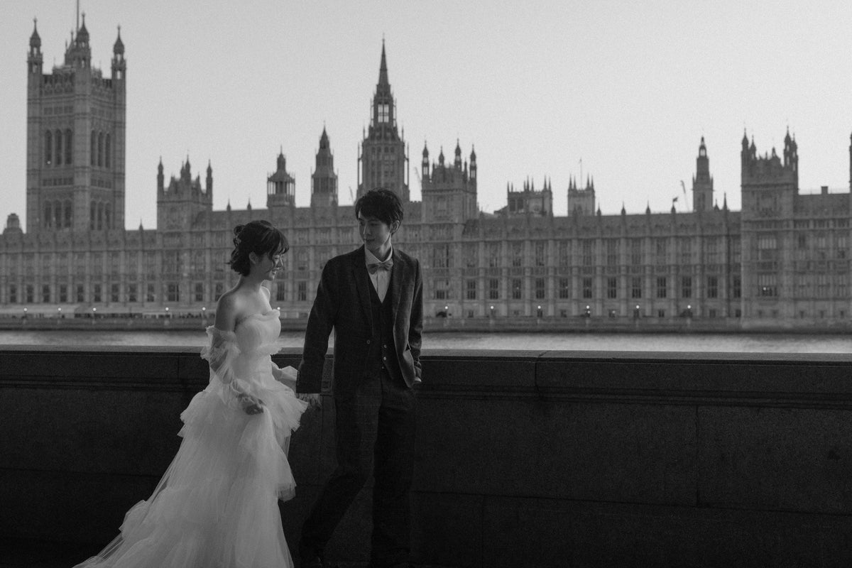 Black and white wedding photograph showing a bride in a flowing white gown and a groom in a dark tuxedo standing in front of a large historic building.