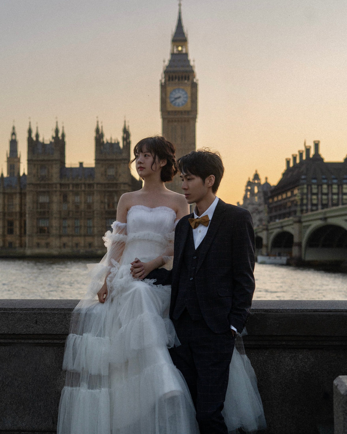 A couple posing in front of London's Big Ben and Westminster Bridge at sunset. The woman wears a strapless white tulle wedding gown, and the man wears a dark three-piece suit with a brown bow tie.