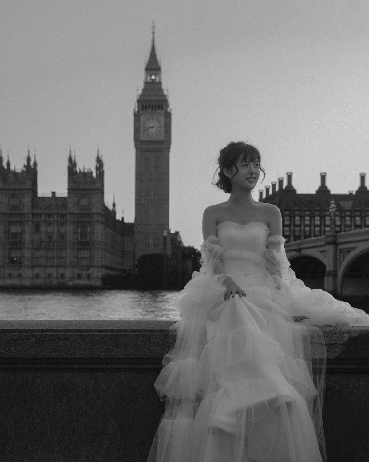 A woman in a strapless white tulle wedding gown with long sheer sleeves stands by a bridge in front of Big Ben and the Houses of Parliament, shown in black and white.