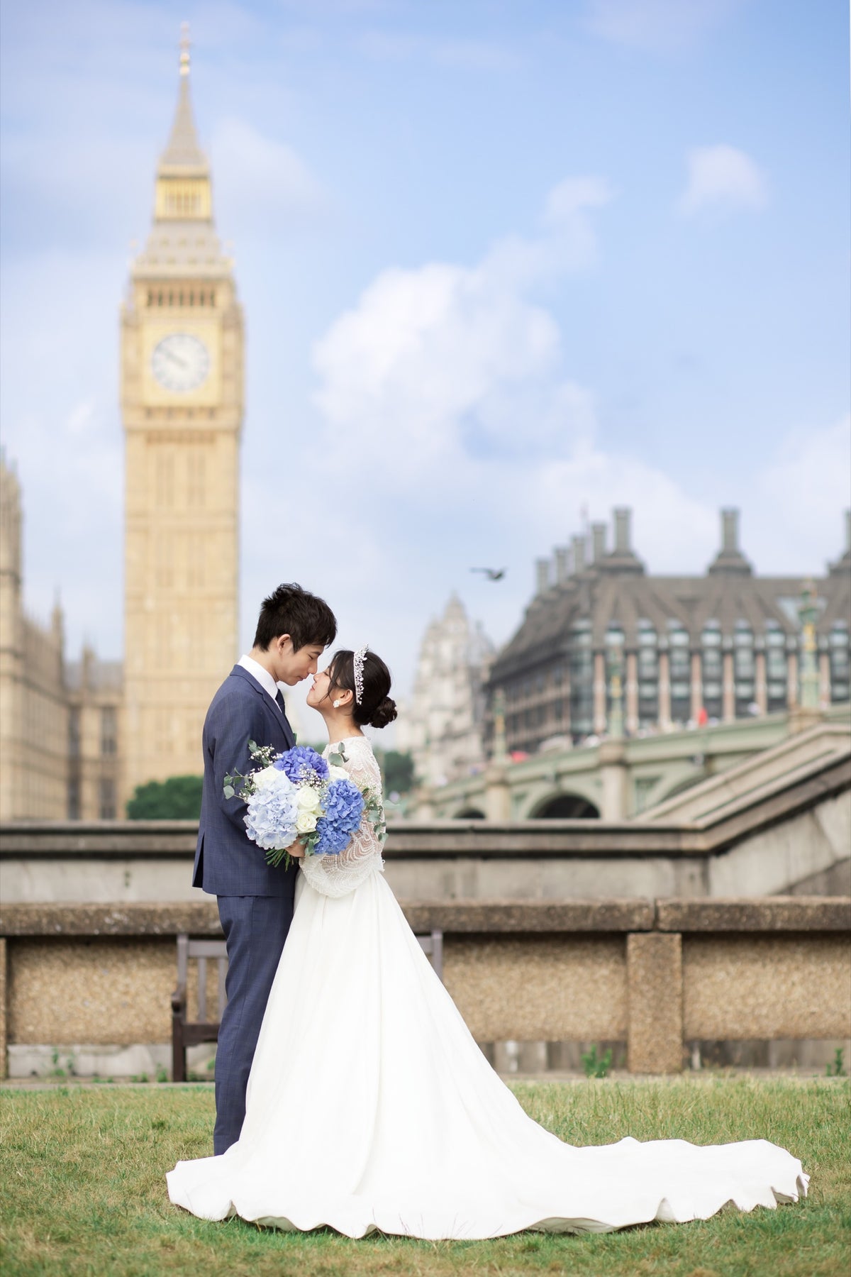 A bride in a white wedding gown and a groom in a navy suit stand together on grass in front of Big Ben and the London Eye under a blue sky with clouds.