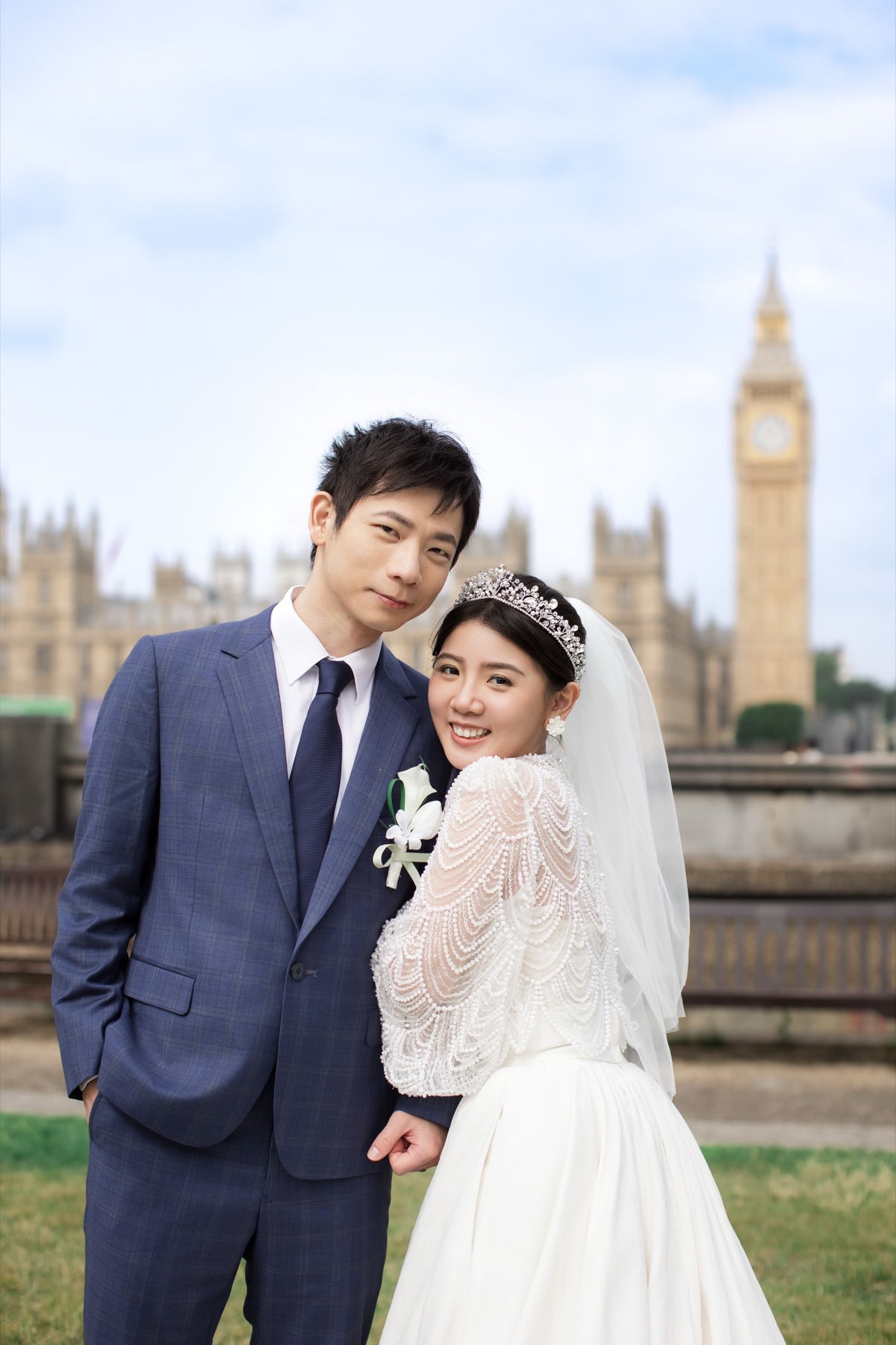 A bride in a white lace wedding gown and veil with a tiara stands beside a groom in a navy blue suit and tie, posing in front of Big Ben and the London Eye under a cloudy sky.