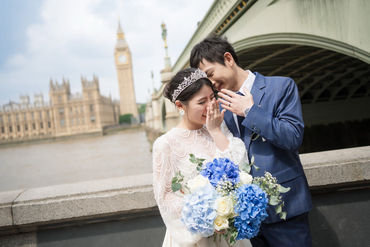 A bride in a white lace wedding gown and tiara embraces a groom in a navy blue suit, holding a bouquet of blue and white flowers, with Big Ben and Westminster Bridge in London in the background.