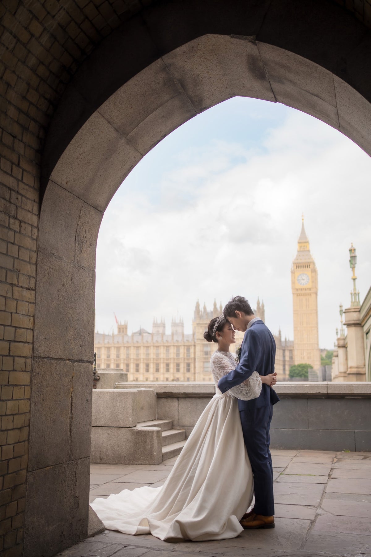 A bride in a white wedding gown and a groom in a navy suit embrace under a stone archway, with Big Ben and the Houses of Parliament visible in the background.