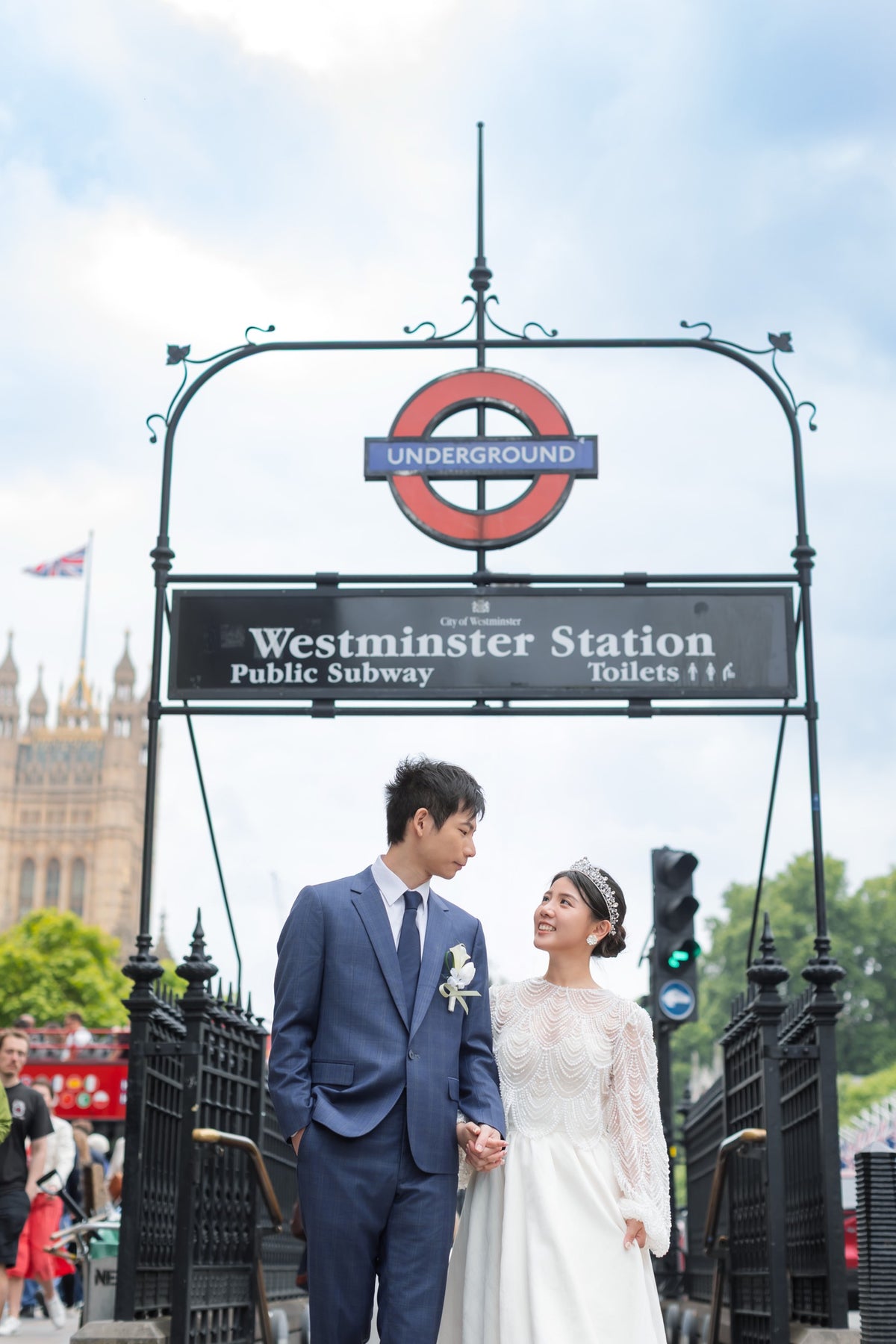 A bride in a white lace wedding gown and a groom in a navy blue suit stand holding hands in front of a Westminster Station Underground roundel sign, with London landmarks and a red double-decker bus in the background.