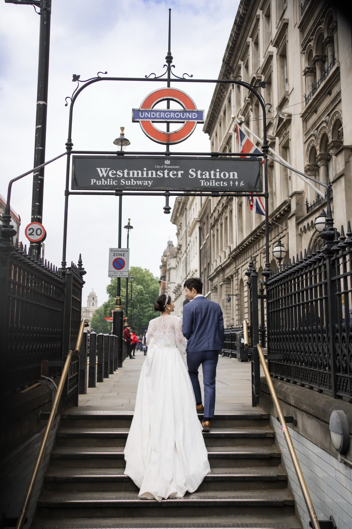 A bride in a white wedding gown and a groom in a navy suit walk up the stairs toward Westminster Station Underground station in London, framed by the station sign and historic stone buildings.