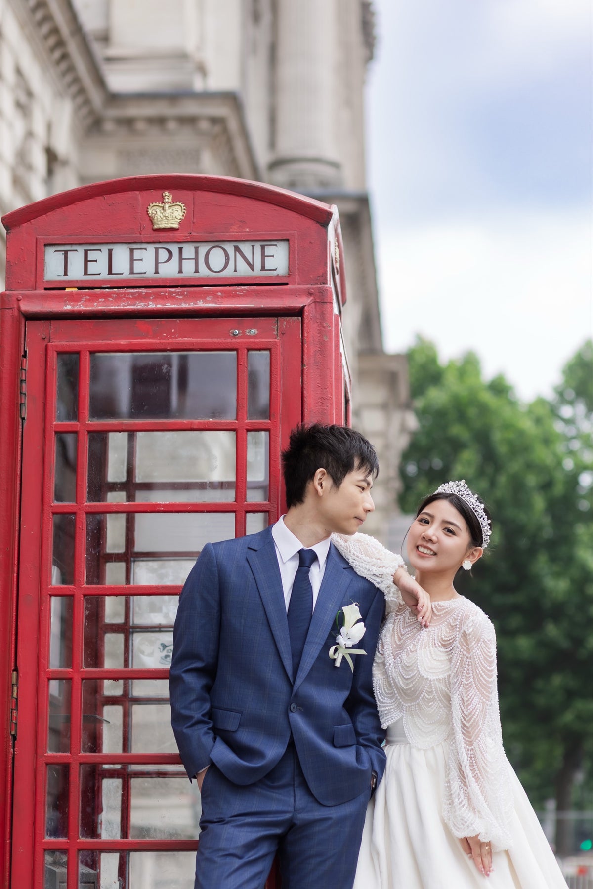 A bride in a white lace wedding gown and a groom in a navy blue suit stand smiling next to a classic red London telephone booth, with a stone building and green trees in the background.