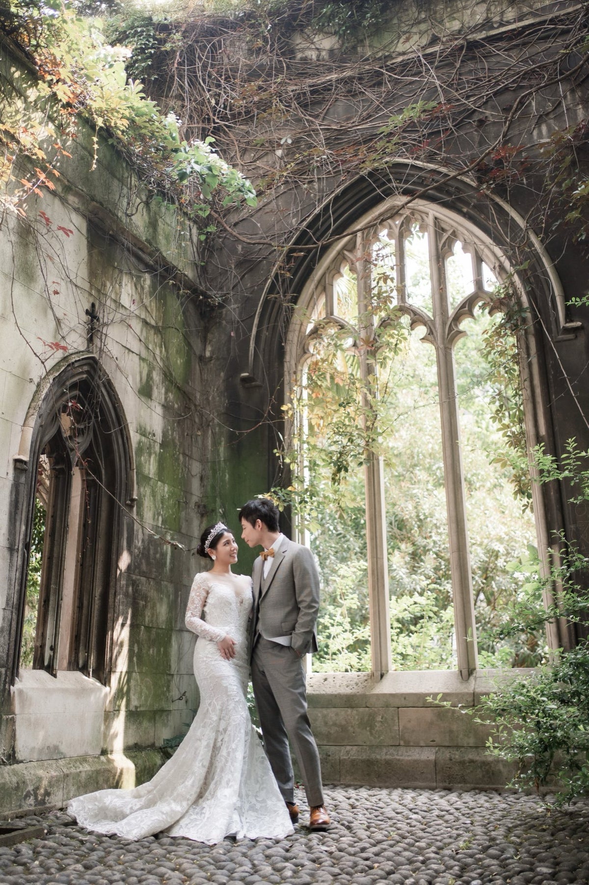 A bride in a white lace wedding gown and a groom in a gray suit stand together in a historic stone ruin with large arched windows and climbing vines.