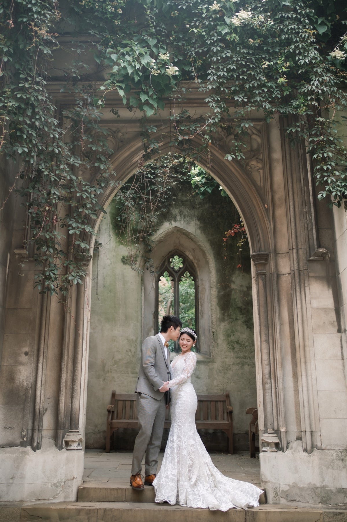 A bride in a white lace wedding gown and a groom in a gray suit pose together in front of a historic stone gothic archway covered in green ivy.