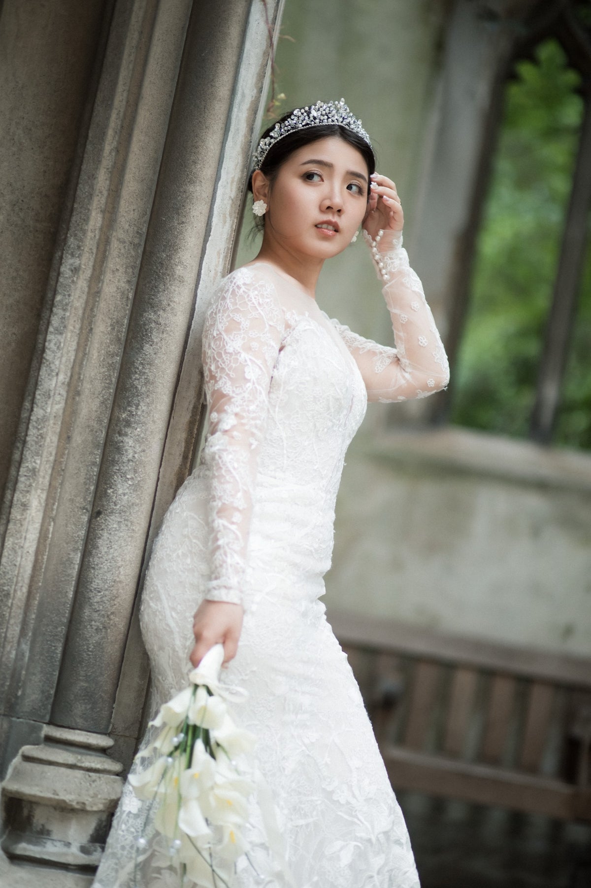 A woman wearing a white lace wedding dress, tiara, and earrings stands outdoors against a stone architectural background, holding a bouquet of white flowers.