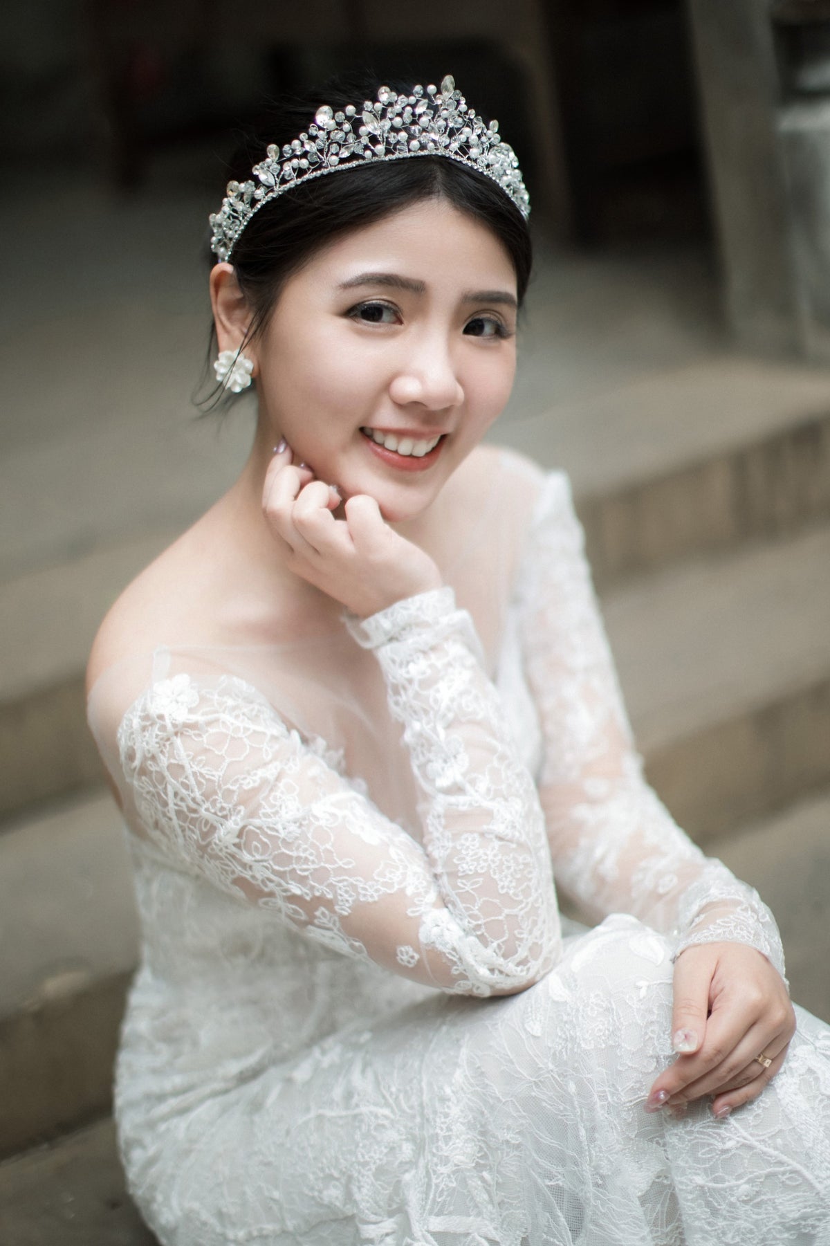A smiling woman in a white lace wedding gown and jeweled tiara sits on stone steps, with her hand near her face.