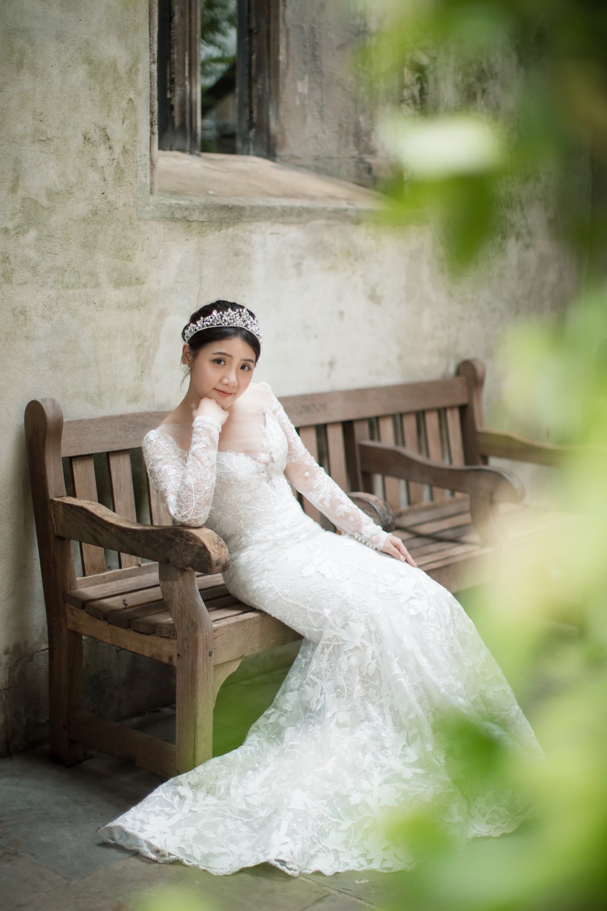 A woman in a white lace wedding gown and tiara sits on a wooden bench against a textured wall, framed by soft green foliage in the foreground.