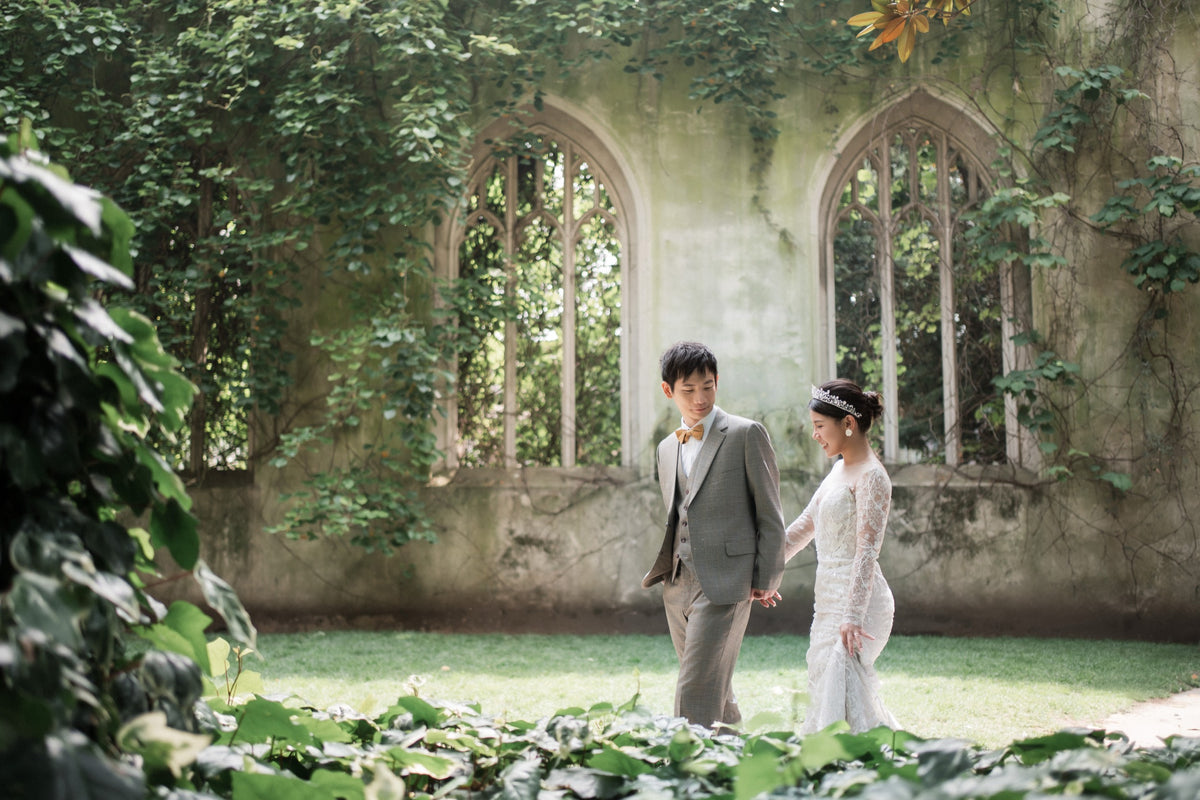 A bride in a white lace wedding gown and a groom in a gray suit holding hands, walking in a grassy garden courtyard with a stone wall and arched windows covered in green ivy behind them.