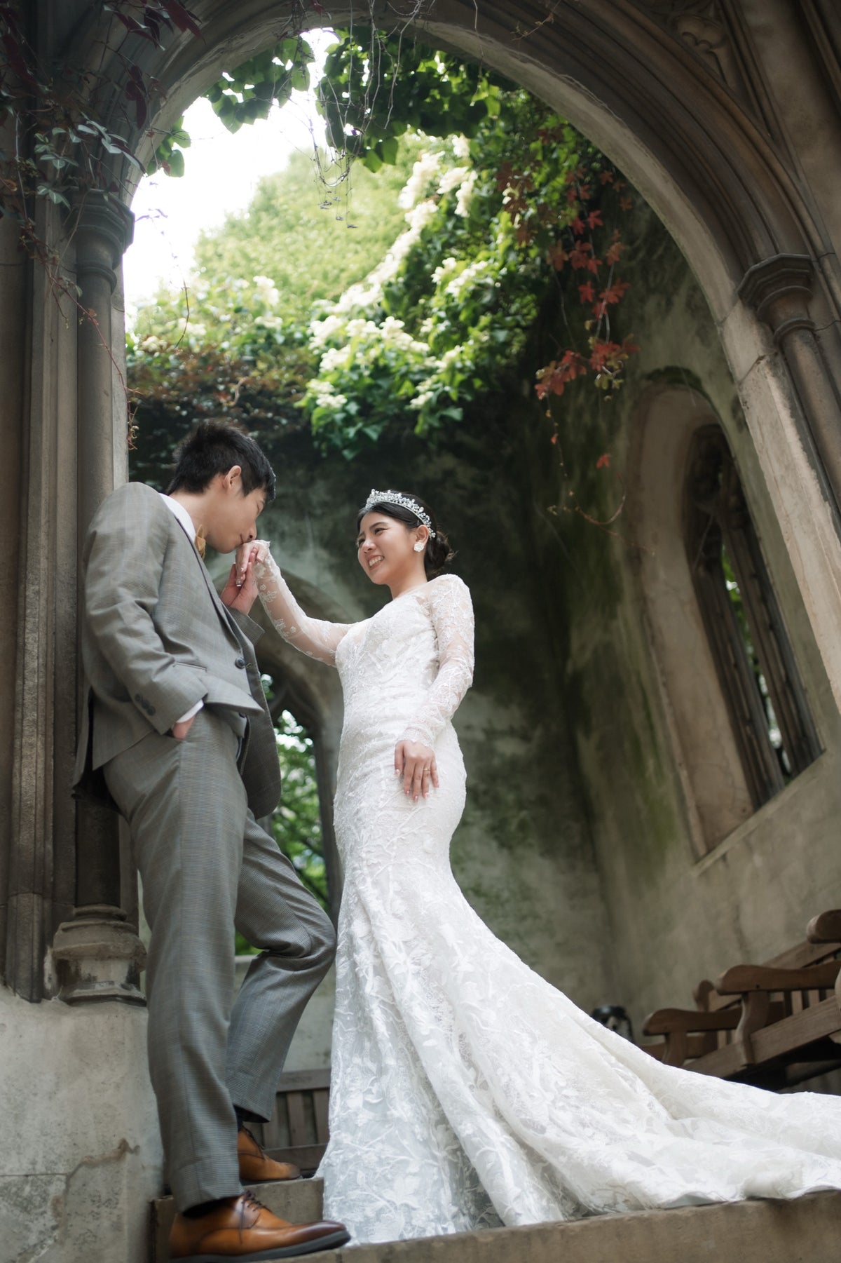 A bride in a white lace wedding gown and a groom in a gray suit pose together on stone steps framed by an arched stone doorway with green foliage above.