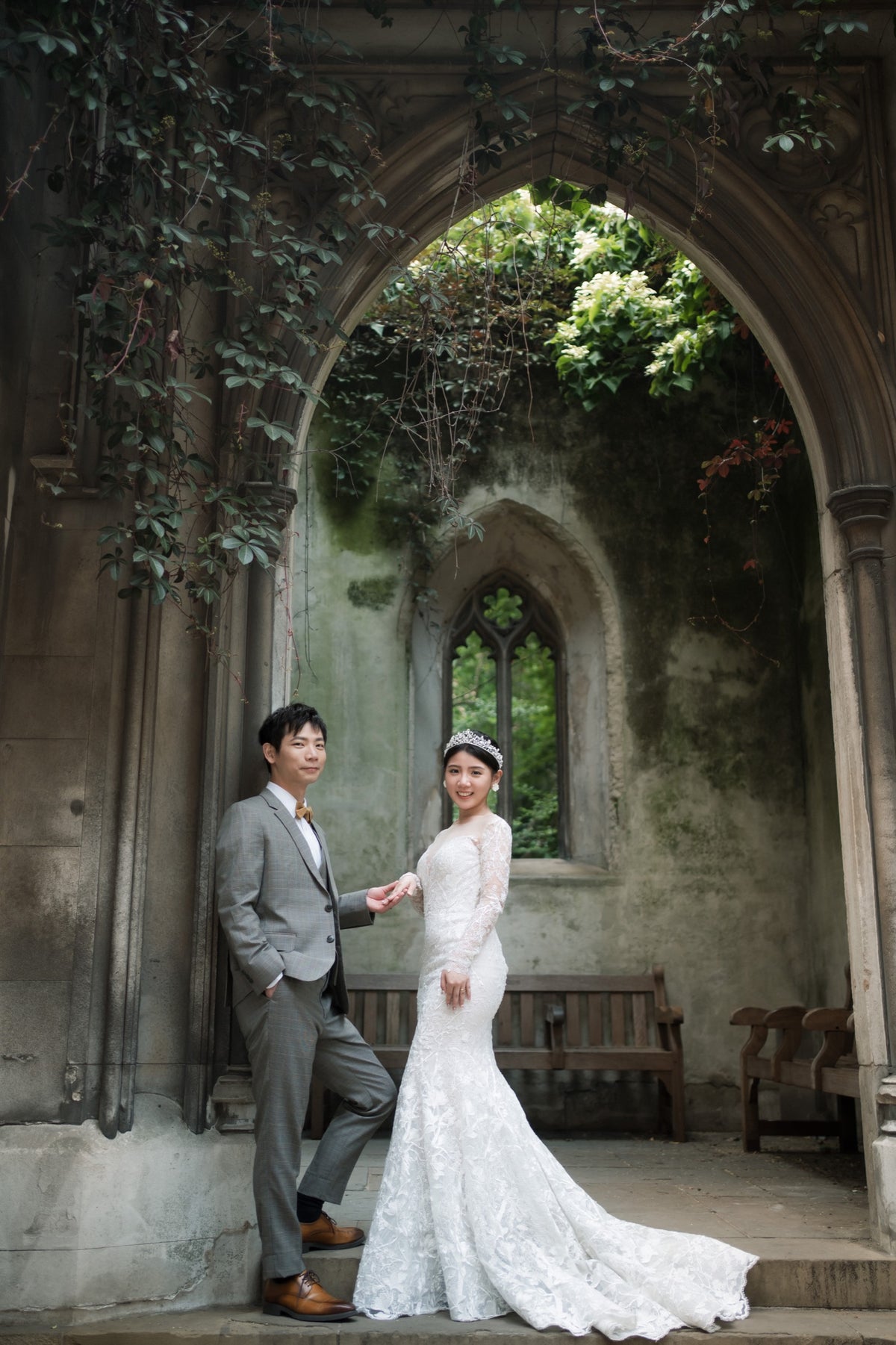 A bride in a white lace wedding gown and a groom in a gray suit pose together inside a historic stone archway with ivy-covered walls and a bench in the background.