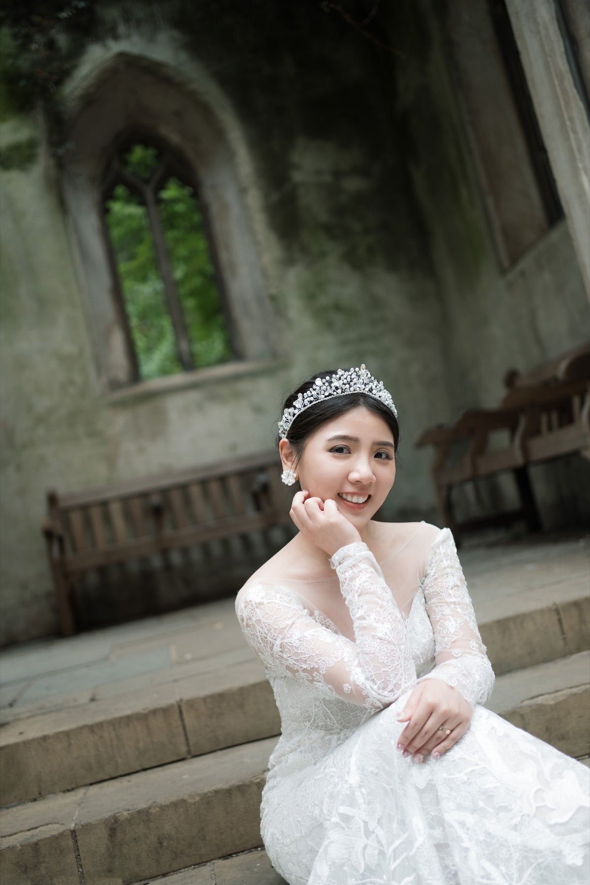 A woman wearing a white lace wedding dress and a sparkling tiara sits on stone steps in front of a stone building with arched windows.