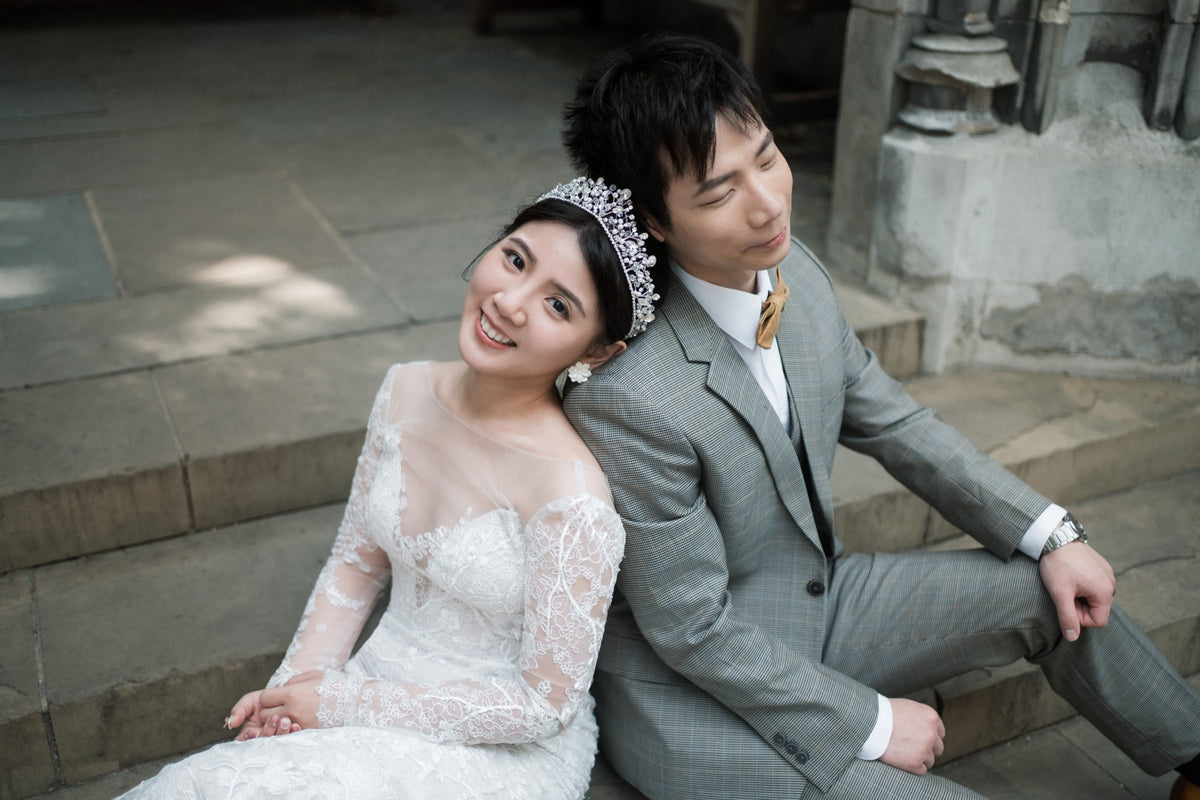 A smiling bride in a white lace wedding gown and tiara sits on stone steps next to a groom in a gray suit and yellow tie.