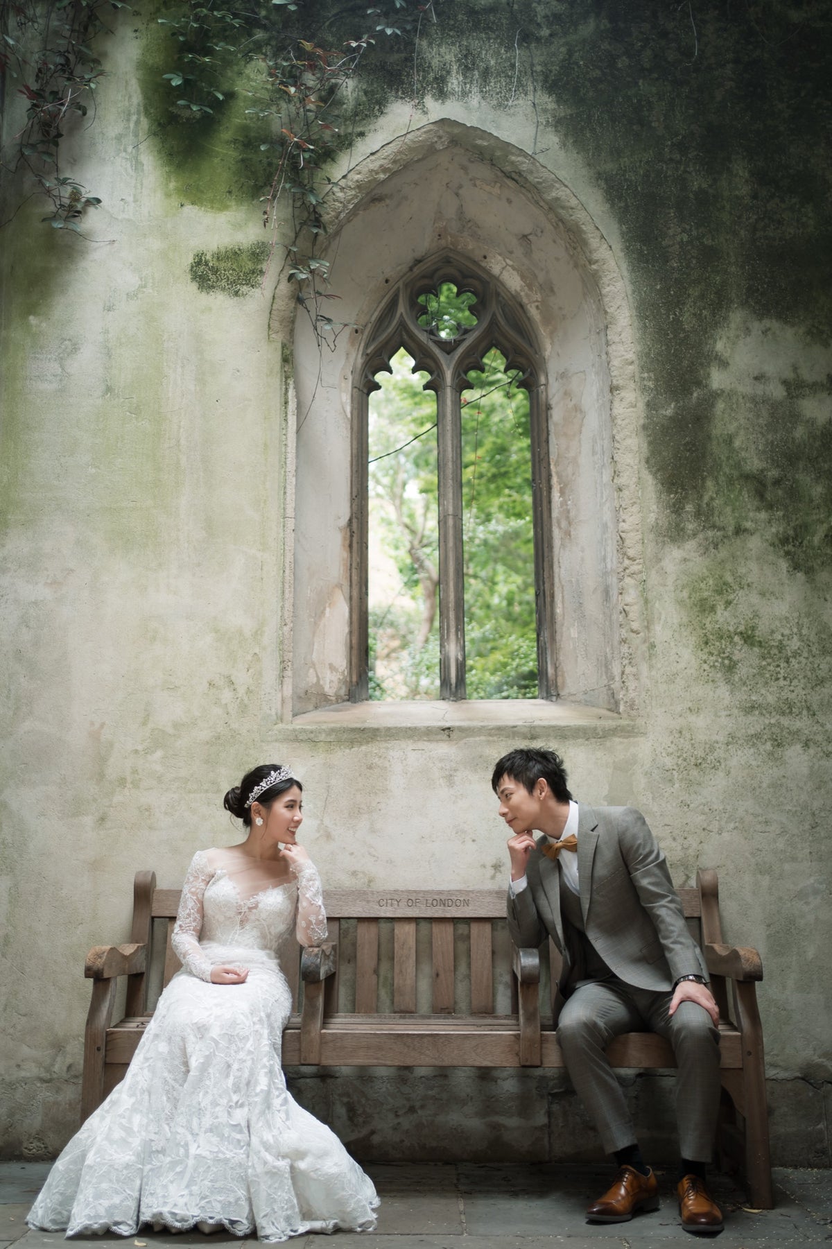 A bride in a white lace wedding gown and a groom in a gray suit sit on a wooden bench in front of a weathered stone wall with a tall arched window.