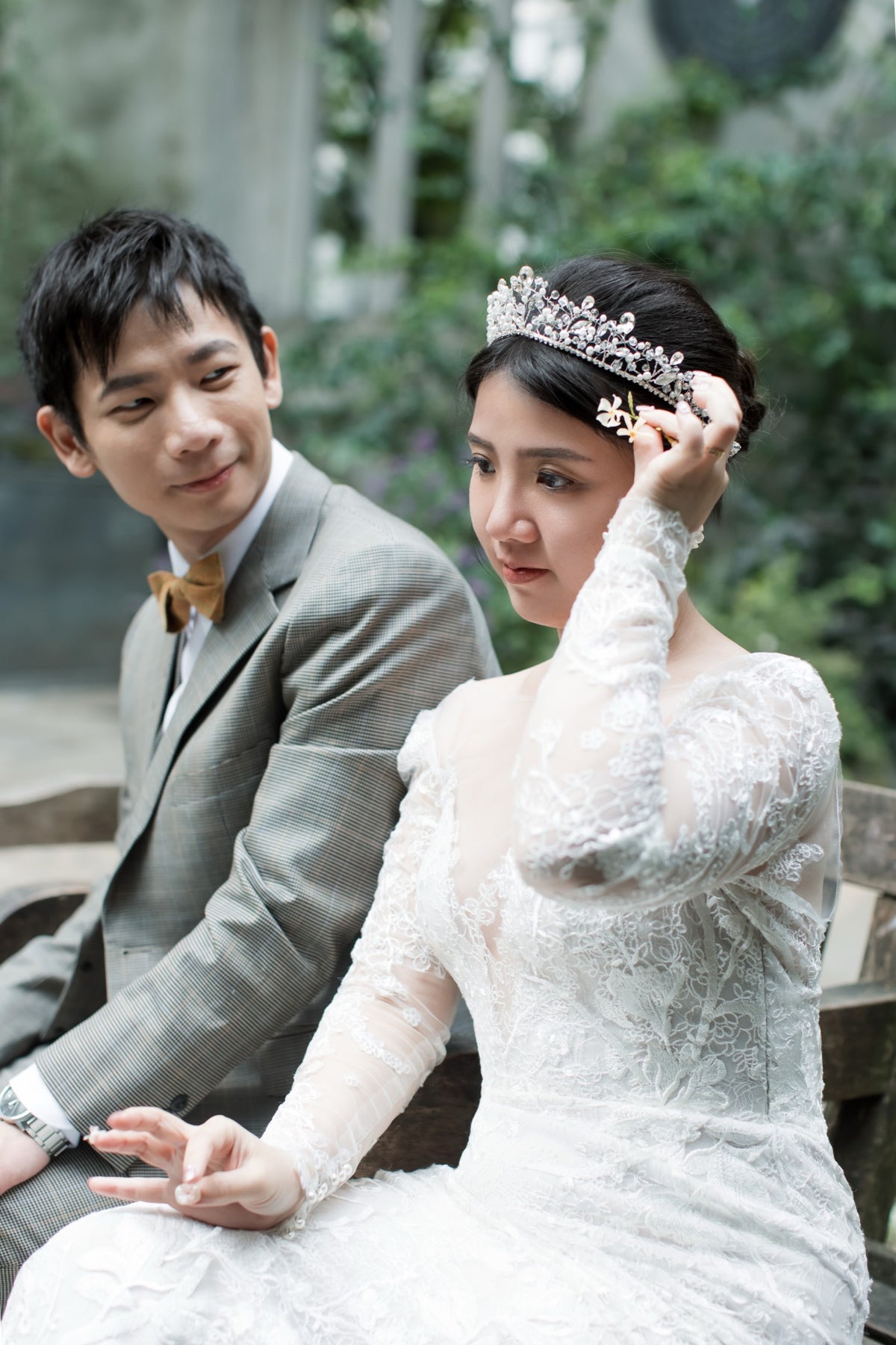 A bride in a white lace wedding gown and tiara sits beside a groom in a gray suit on a wooden bench, with a softly blurred outdoor garden background.