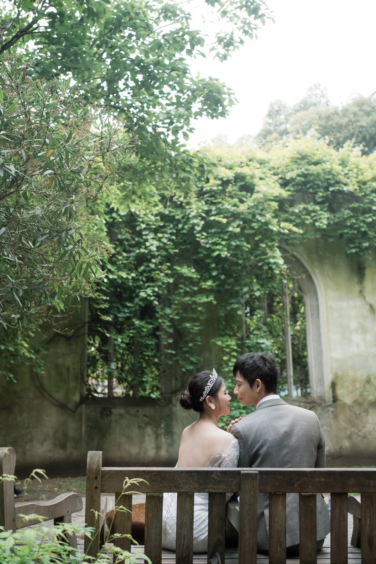 A couple in wedding attire sits on a wooden bench in a garden setting with lush green ivy-covered walls and trees.