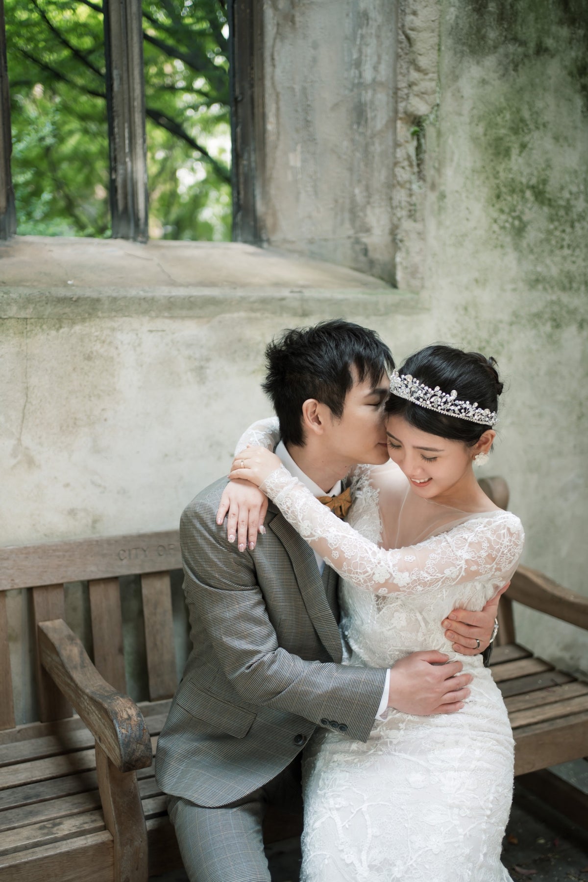 A bride in a white lace wedding gown and tiara embraces a groom in a gray suit on a wooden bench against a rustic stone wall.