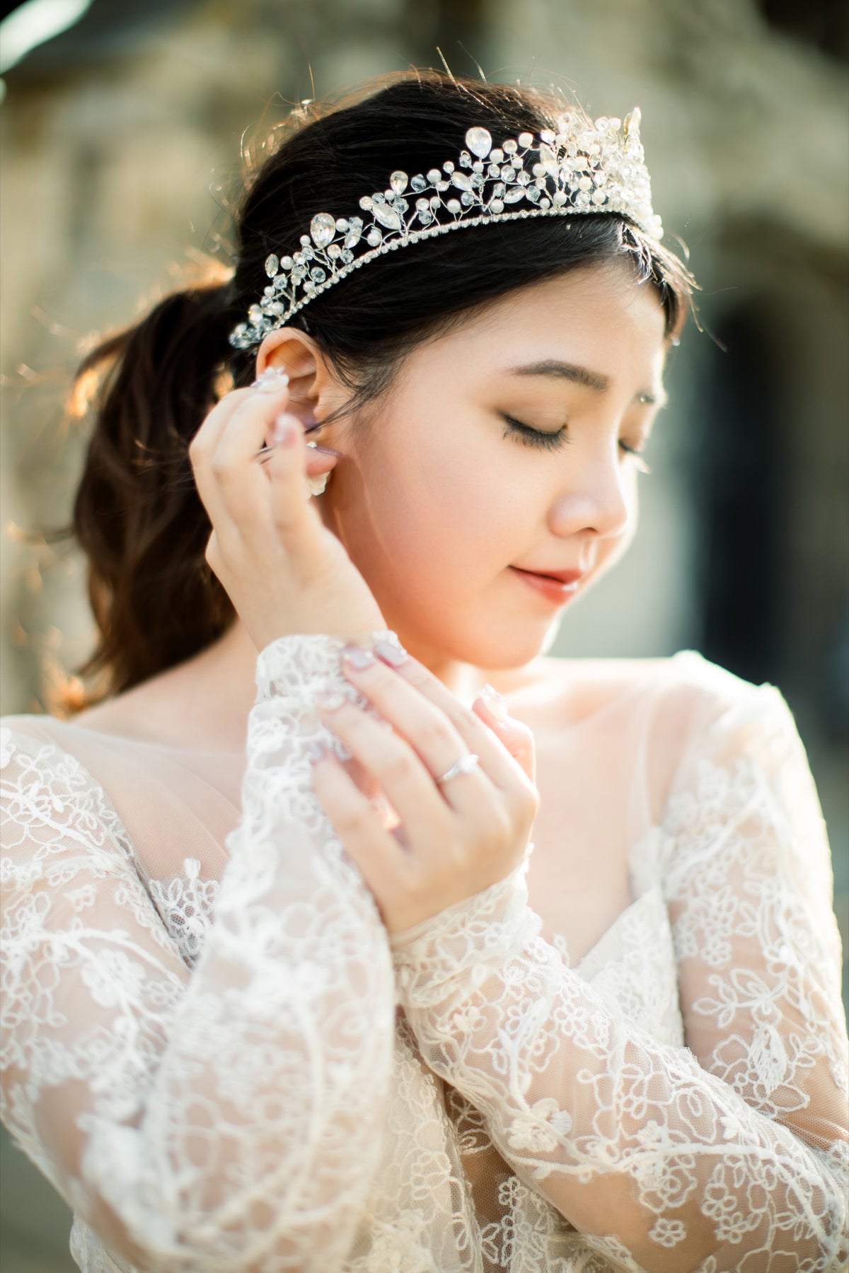 A bride wearing a sparkling silver tiara and white lace wedding dress, adjusting her earring with a soft, warm outdoor background.