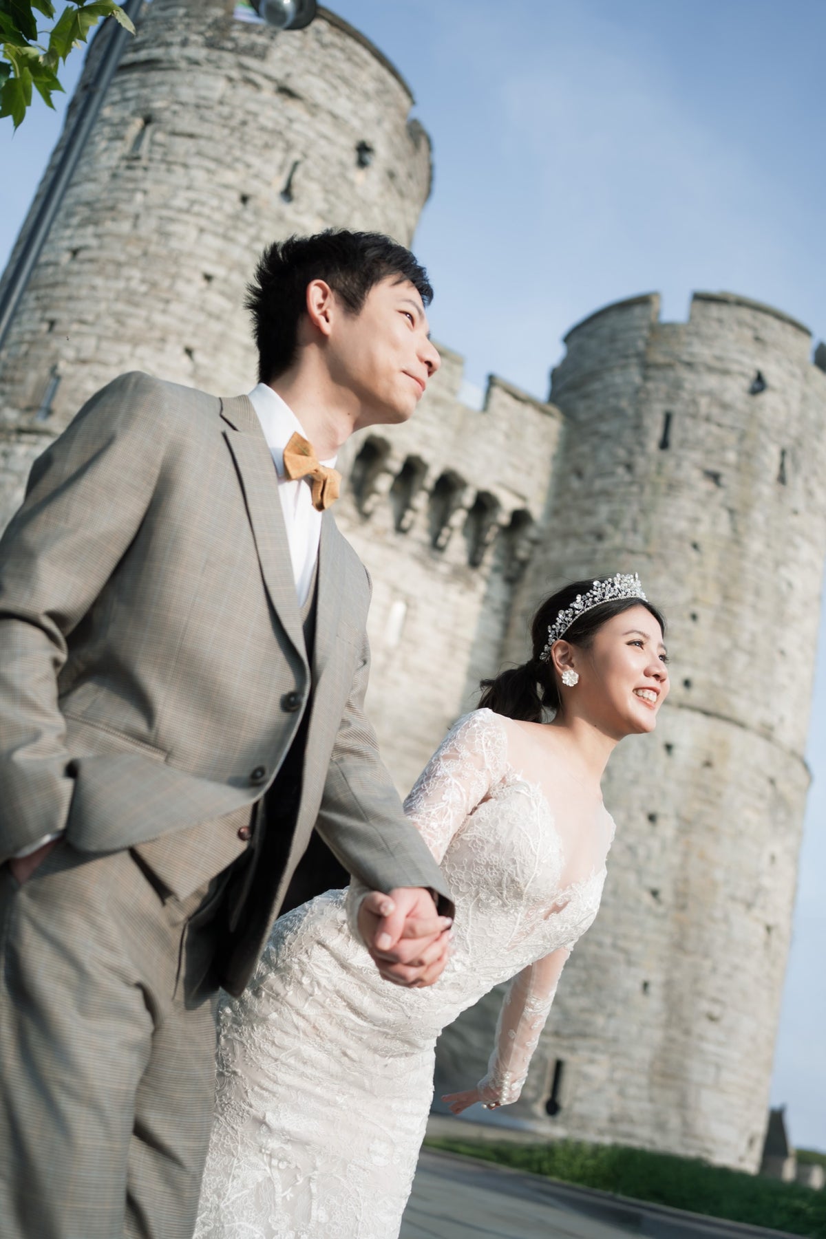 A bride in a white lace wedding gown and tiara walks hand-in-hand with a groom in a light gray suit and yellow bow tie, photographed outdoors in front of a stone castle under a blue sky.