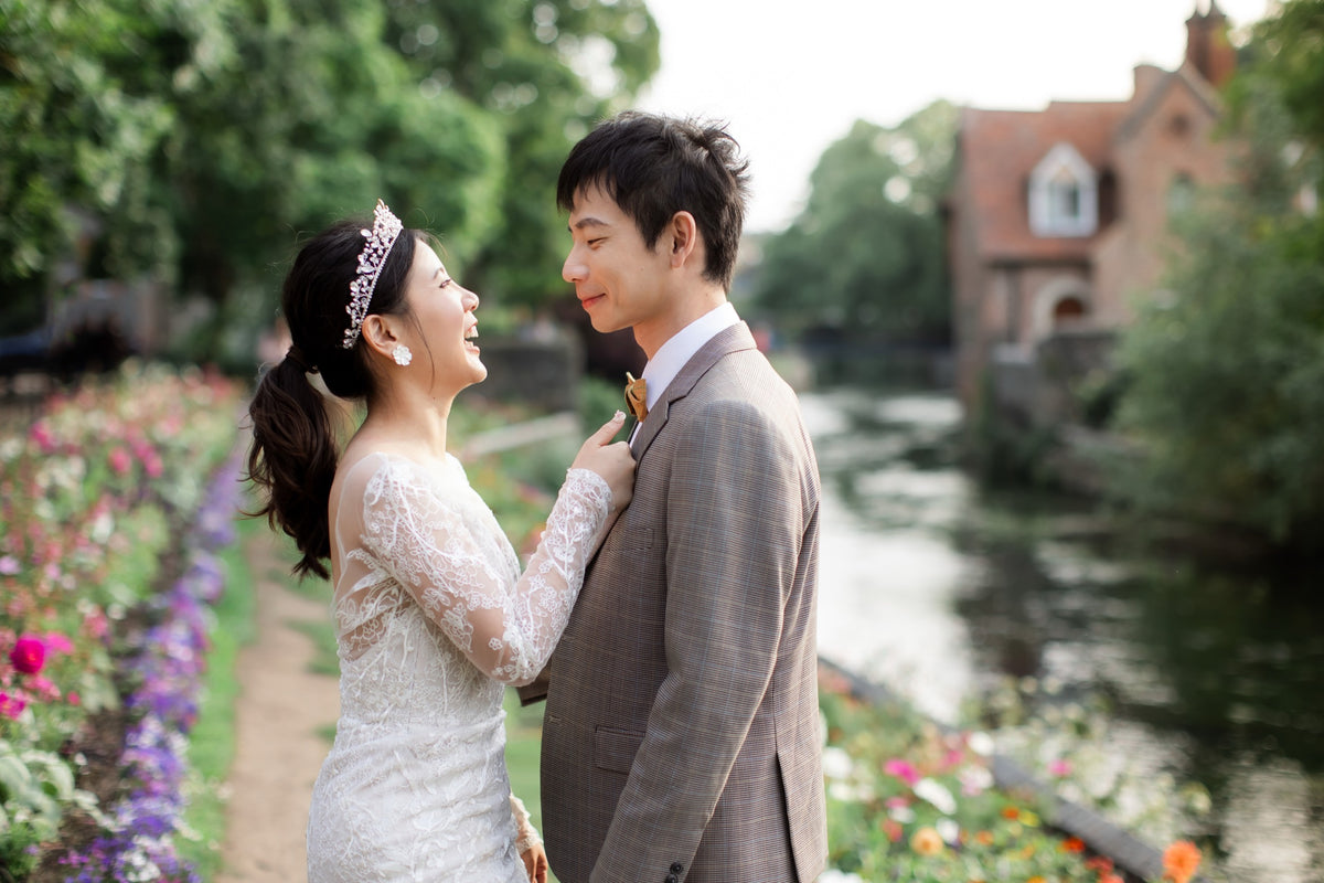 A bride in a white lace wedding dress and tiara smiles at a groom wearing a gray suit, standing by a canal with flowers and a building in the background.