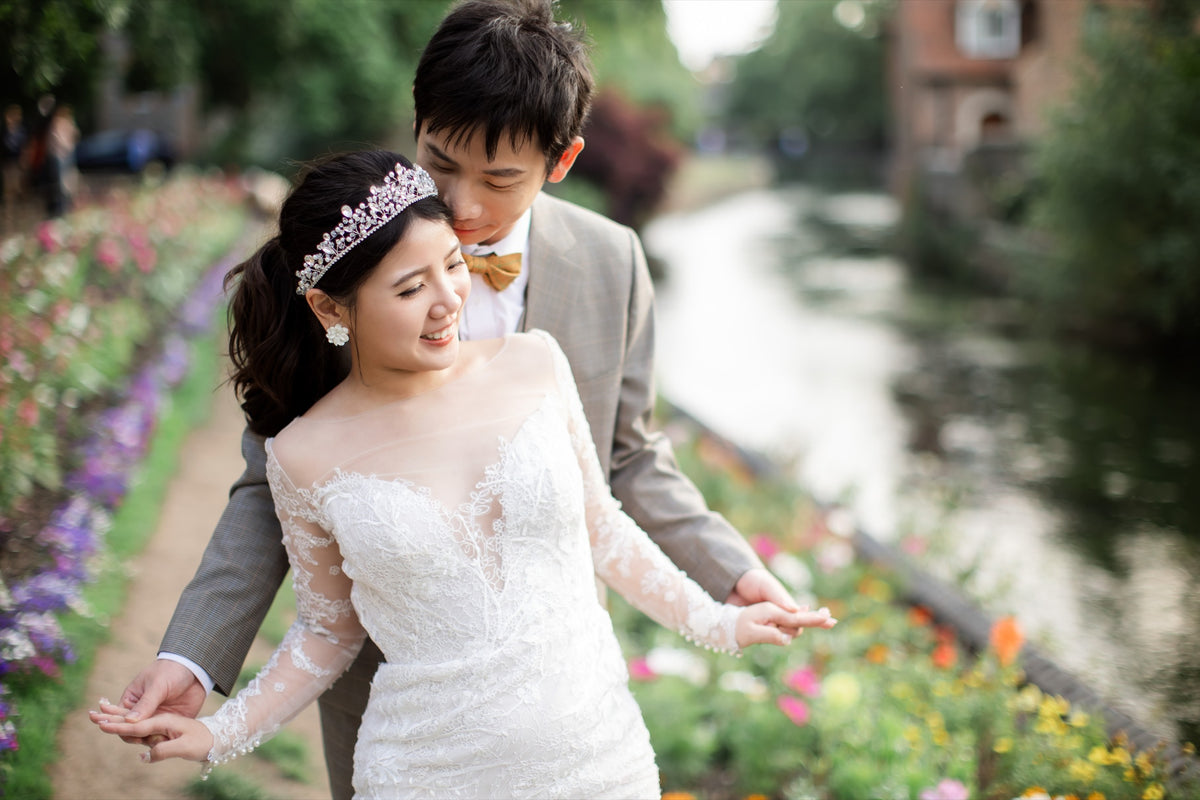 A bride in a white lace off-the-shoulder wedding dress with a sparkling tiara walks hand in hand with a groom in a light gray suit and yellow bow tie along a canal path lined with flowers.