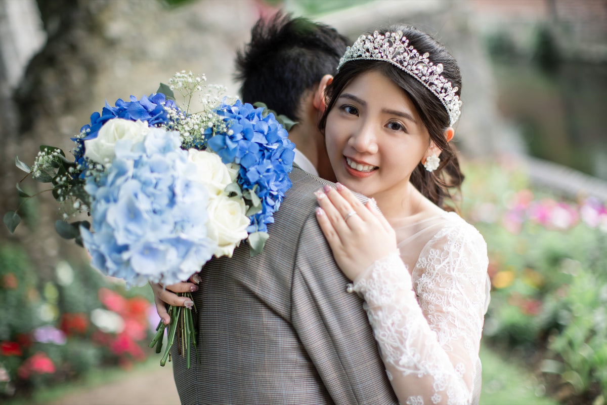 A smiling bride in a white lace wedding dress and tiara hugs a man outdoors, holding a bouquet of blue and white flowers. The background is a softly blurred garden setting.