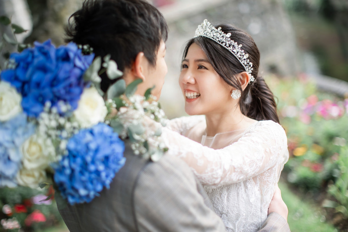 A smiling bride in a white lace wedding dress and silver tiara embraces a groom holding a large bouquet of blue, white, and green flowers, set against a softly blurred garden background.