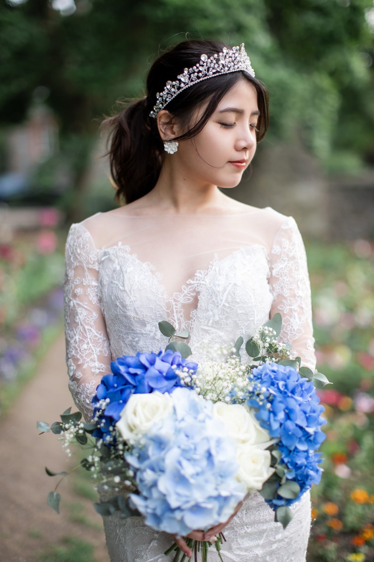A bride in a white lace wedding gown wearing a sparkling tiara, holding a bouquet of blue and white flowers in a garden setting.